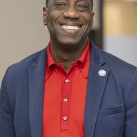 Dr. Julius Sokenu smiling, wearing a red shirt and blue sport coat with an MC Pin