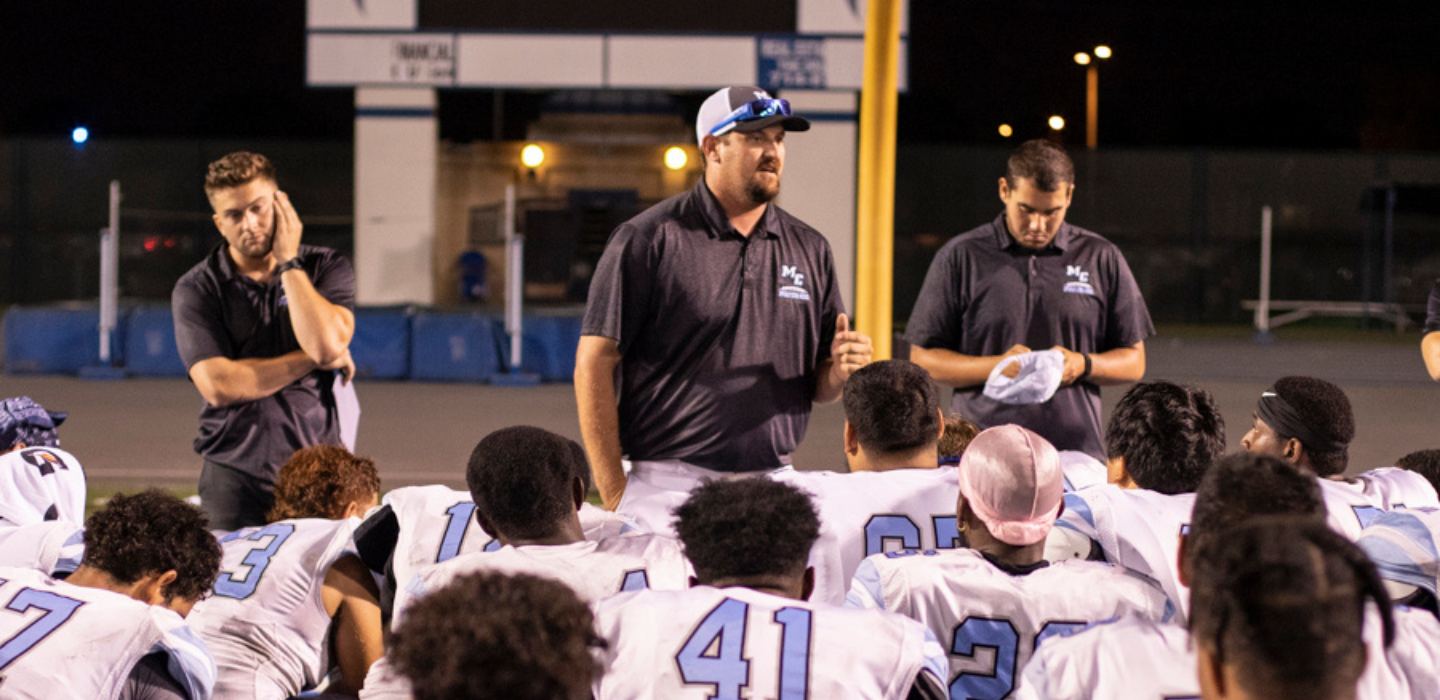 Football team members take a knee while listening to their coach.