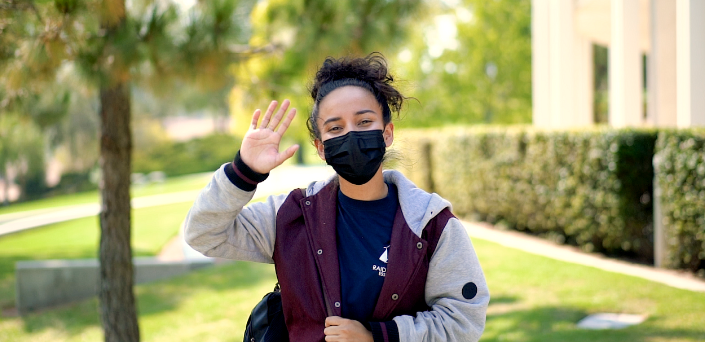 A student raises her hand to the camera while standing outside on campus