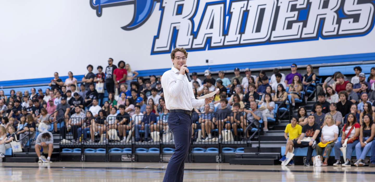 Man standing with microphone in front of crowd sitting in bleachers in Moorpark College gym