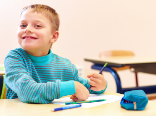 A young child smiling at a school desk while writing.