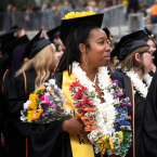 A smiling graduate holds a bouquet and wears colorful leis a
