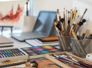 A desk with an assortment of art supplies and a laptop and notebooks.