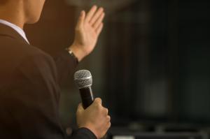 Person speaking with a microphone in one hand and gesturing with the other.