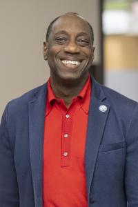 Dr. Julius Sokenu smiling, wearing a red shirt and blue sport coat with an MC Pin